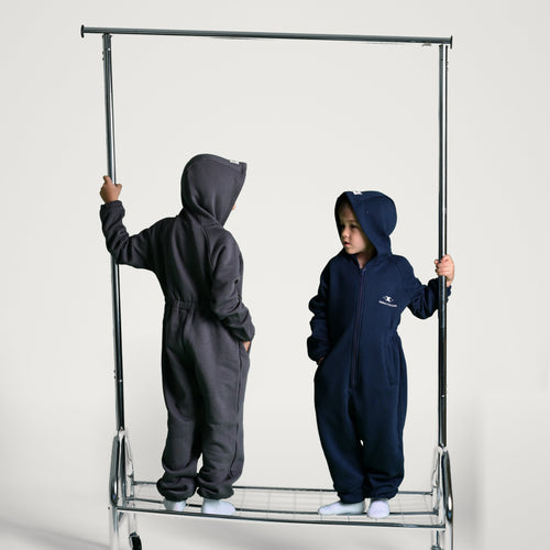 Two children wearing full-body hooded onesie outfits stand on a metal rolling garment rack against a light gray studio background. The child on the left wears a dark charcoal gray hooded jumpsuit, and the child on the right wears a dark navy hooded jumpsuit with a small white chest logo.